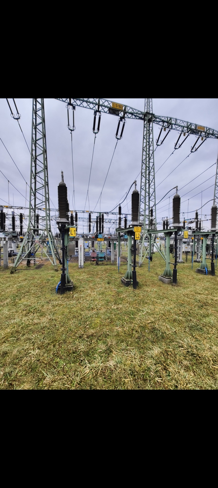 Rozległa stacja transformatorowa z konstrukcjami wsporczymi i izolatorami, widoczna na tle pochmurnego nieba i trawiastym podłożu. Oznaczenia L1, L2, L3 na słupach.
