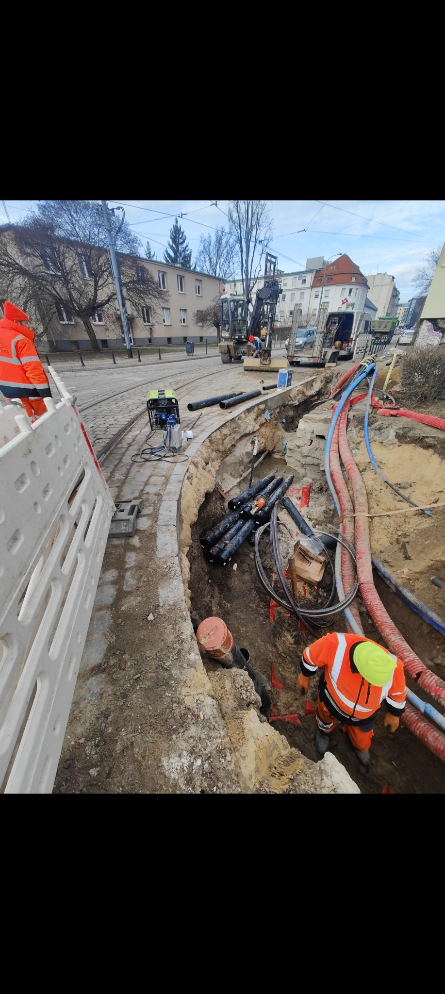 Wykop z widocznymi rurami i pracownikami w odblaskowych ubraniach na tle miejskiej ulicy z torami tramwajowymi. Prace ziemne w toku.