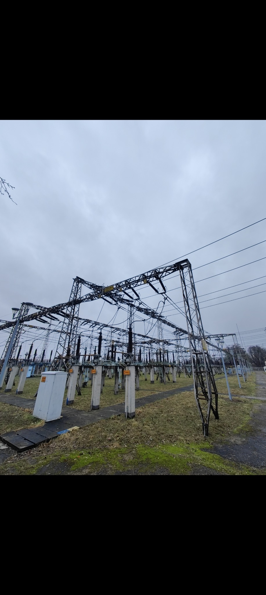Rozległa stacja transformatorowa z metalowymi konstrukcjami i betonowymi podporami pod pochmurnym niebem. Widoczne przewody i urządzenia elektryczne.