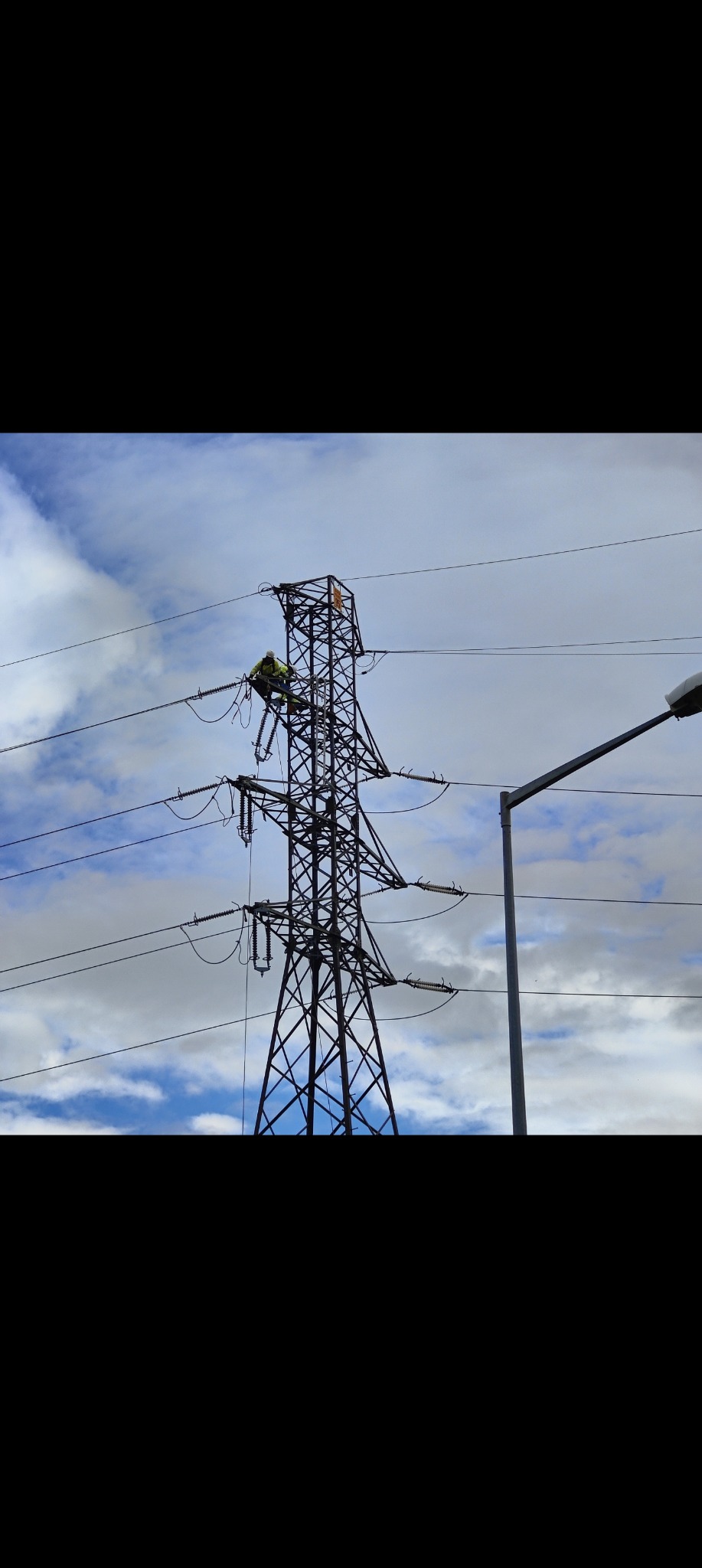Pracownik w kasku na wieży wysokiego napięcia, wykonujący prace konserwacyjne na tle pochmurnego nieba. Widoczny słup oświetleniowy.