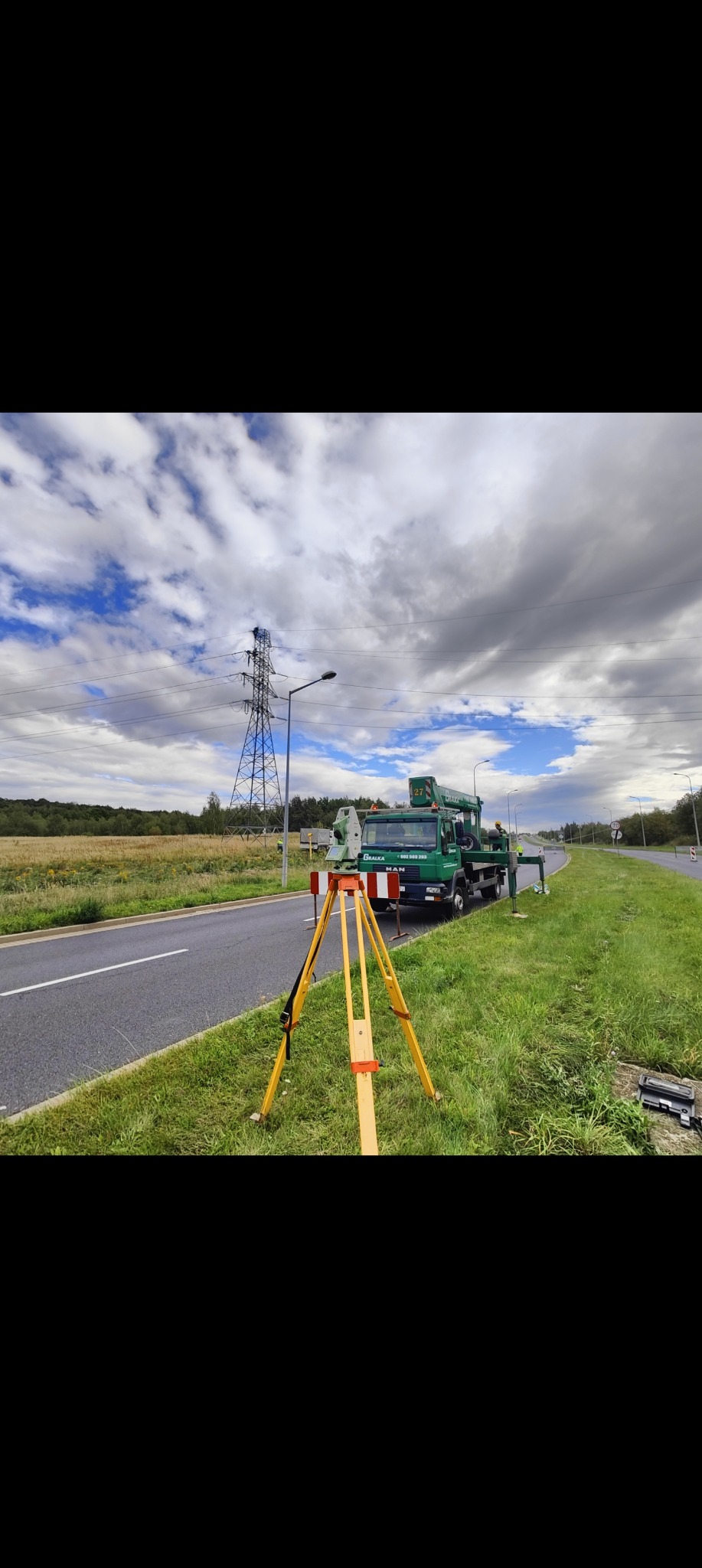 Praca pomiarowa przy drodze: zielony samochód MAN z podnośnikiem, tyczka geodezyjna na trójnogu, w tle słup wysokiego napięcia i pochmurne niebo.