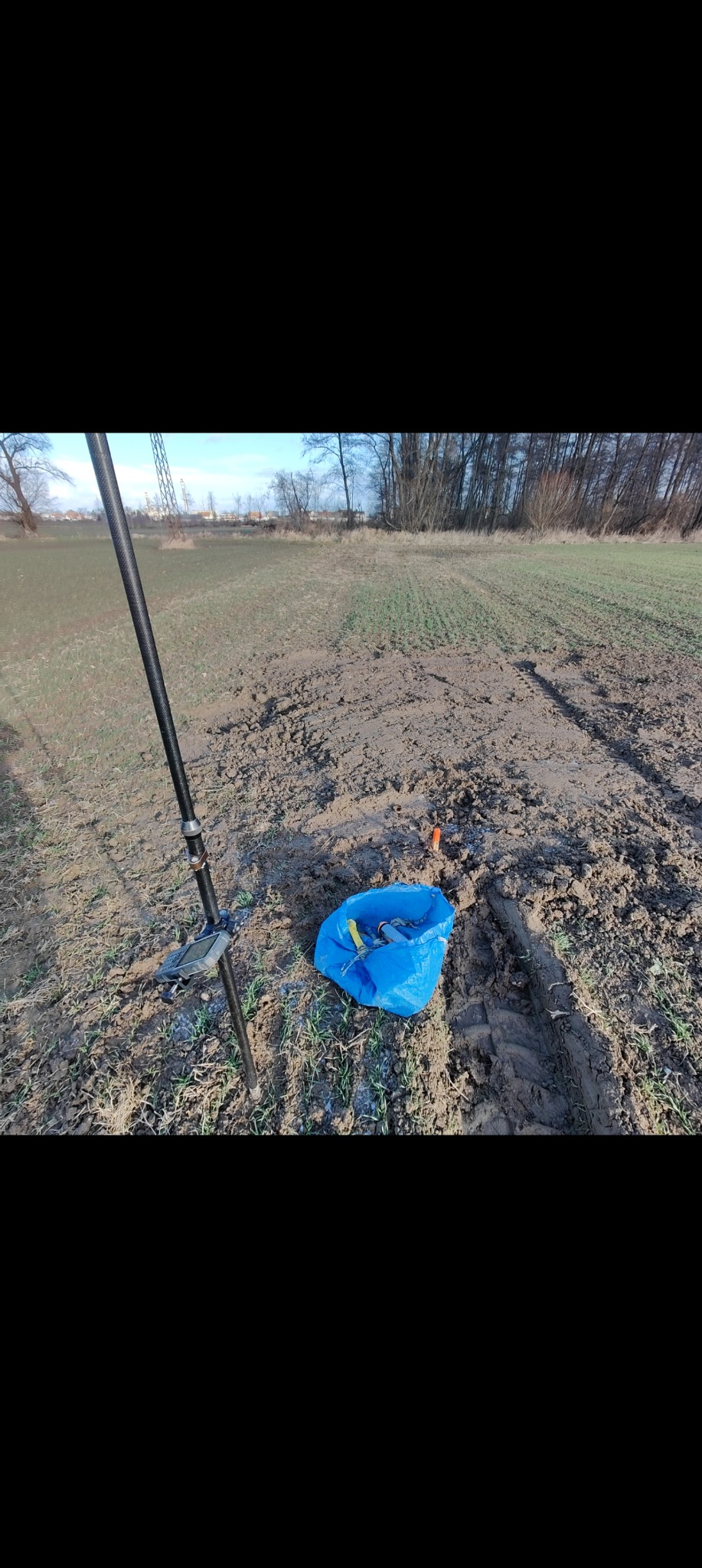 Czarny tyczka geodezyjna wbita w ziemię obok niebieskiej torby z narzędziami na polu. W tle widoczne drzewa i słupy wysokiego napięcia.