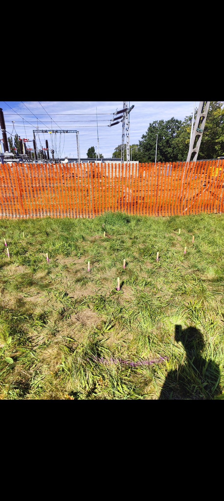 Teren z wyznaczonymi punktami pomiarowymi, zabezpieczony pomarańczową siatką. W tle widoczna infrastruktura energetyczna. Praca w toku.