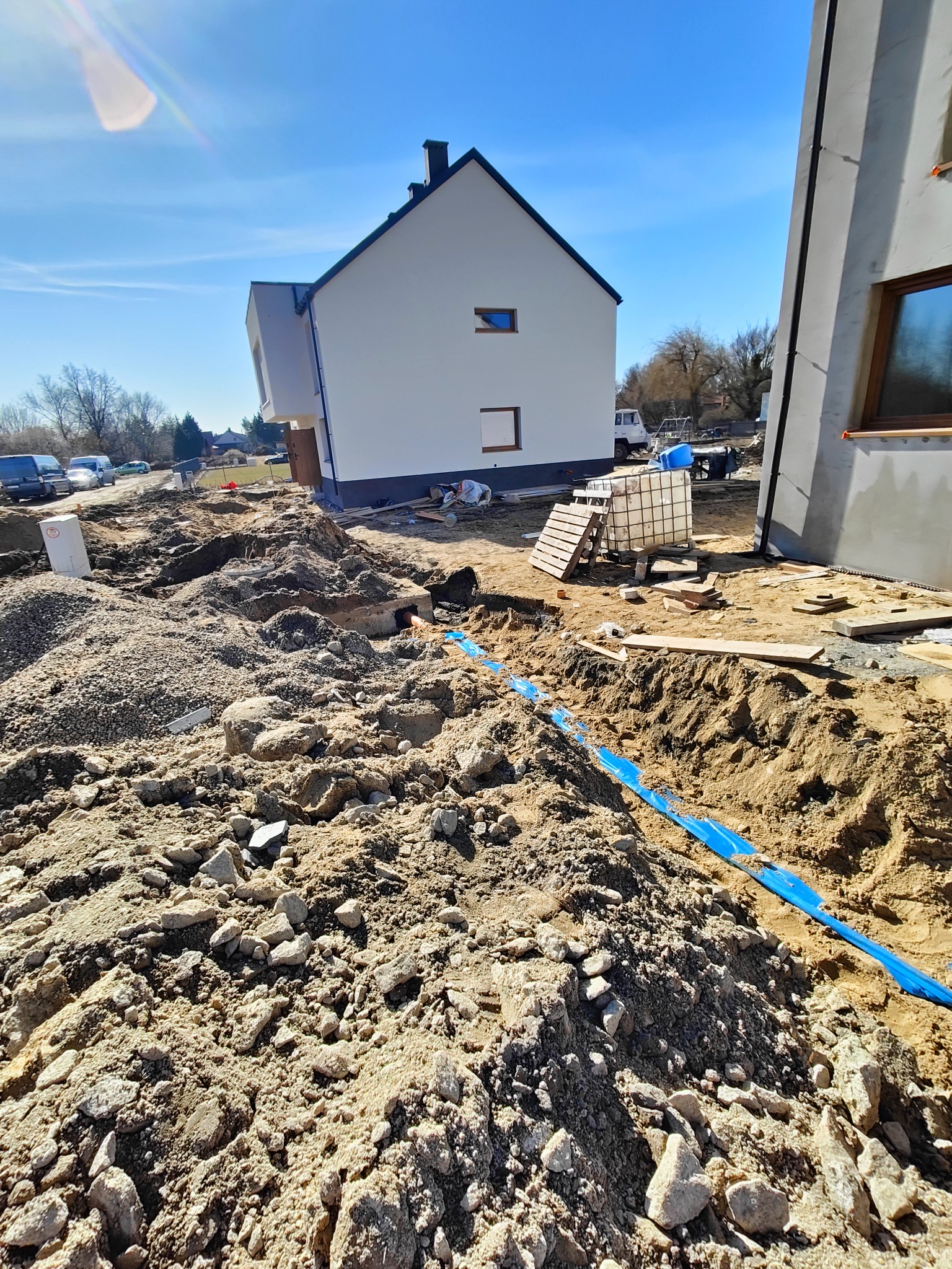 Niebieska rura w wykopie na tle budowy domu jednorodzinnego w słoneczny dzień. Widoczne materiały budowlane i częściowo ukończone budynki.