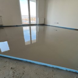 RevoArt - Wylany samopoziomujący podkład podłogowy w jasnym, pustym pokoju z oknem balkonowym. Podkład zabezpieczony niebieską taśmą przy ścianach.