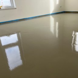 RevoArt - Wylana, gładka posadzka samopoziomująca w jasnym pomieszczeniu z oknem. Niebieska taśma zabezpiecza ściany. Na parapecie leżą narzędzia.