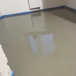 RevoArt - Wylana, gładka wylewka samopoziomująca w pomieszczeniu, zabezpieczona niebieską taśmą malarską przy ścianach. Widoczny grzejnik na ścianie.
