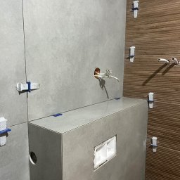 RevoArt - Szare płytki imitujące beton na ścianie i zabudowie WC, z systemem poziomowania płytek. Brązowe płytki drewnopodobne w tle. Widoczne otwory na instalacje.