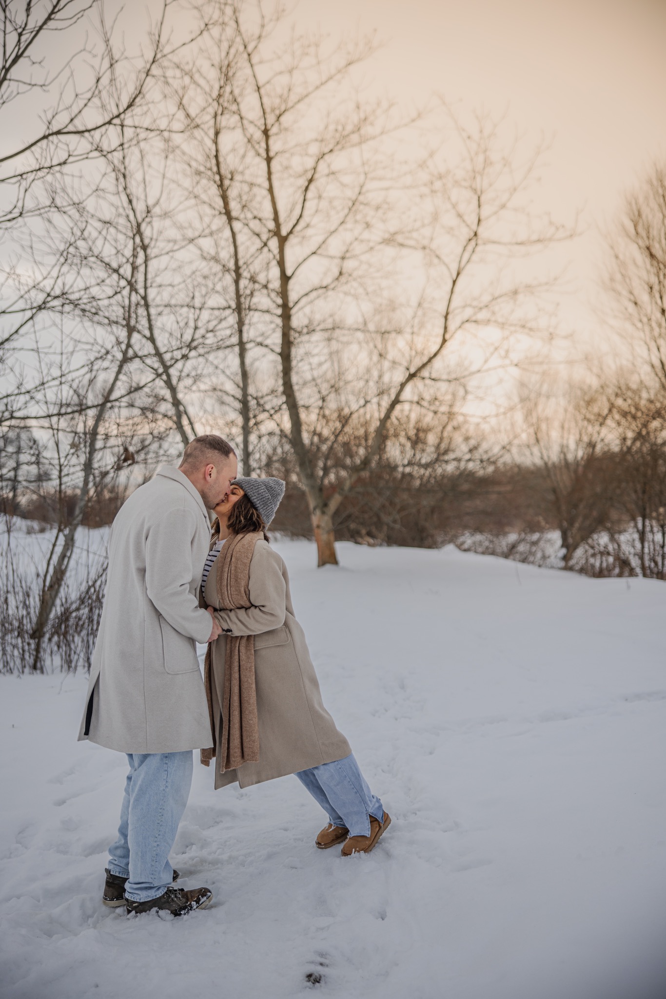 Zimowa sesja narzeczeńska: para w jasnych płaszczach całuje się na tle ośnieżonego krajobrazu z drzewami bez liści, ciepłe, naturalne światło.