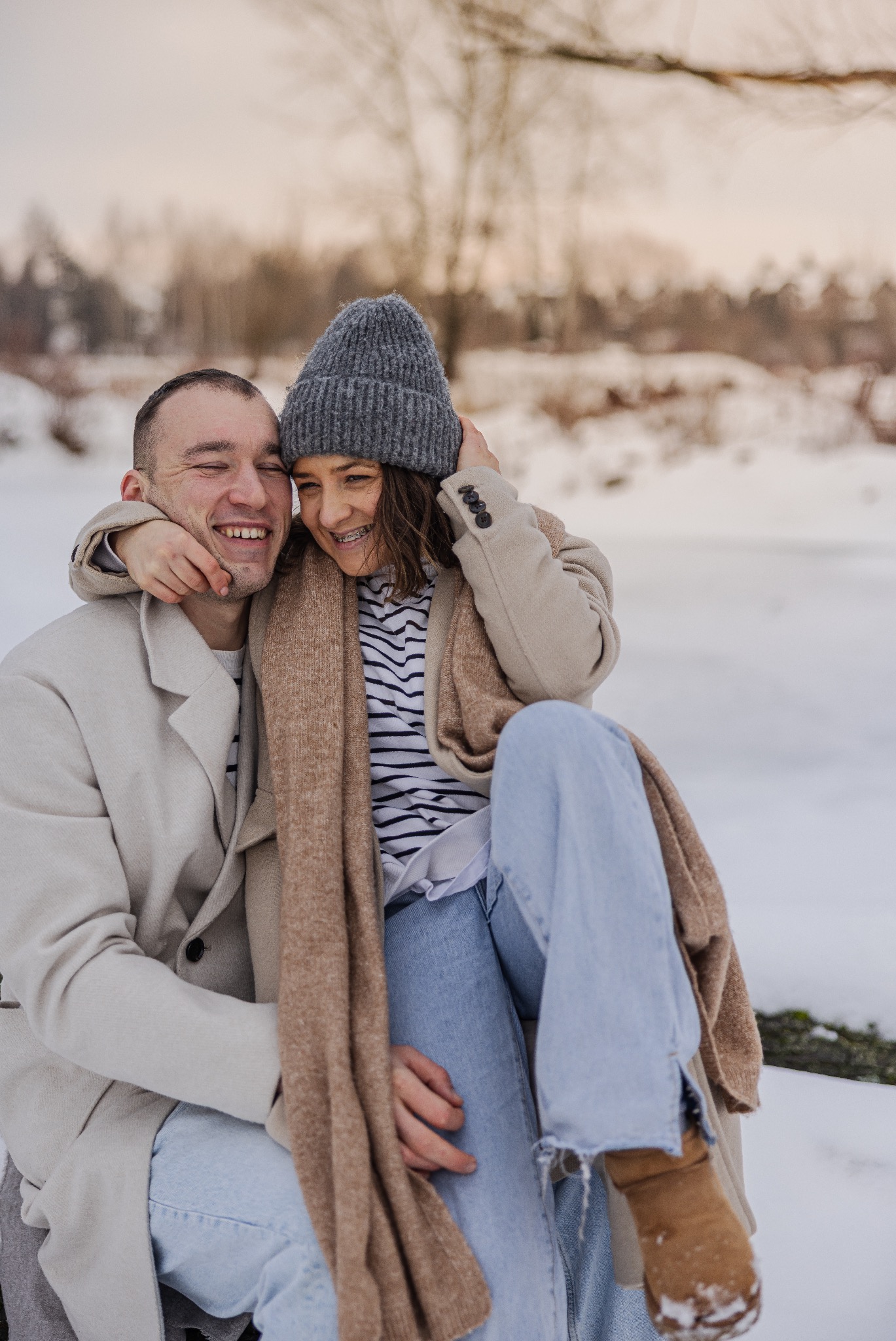 Uśmiechnięta para w zimowej scenerii: mężczyzna obejmuje kobietę w czapce i szaliku, na tle ośnieżonych drzew. Widoczne aparaty ortodontyczne u kobiety.