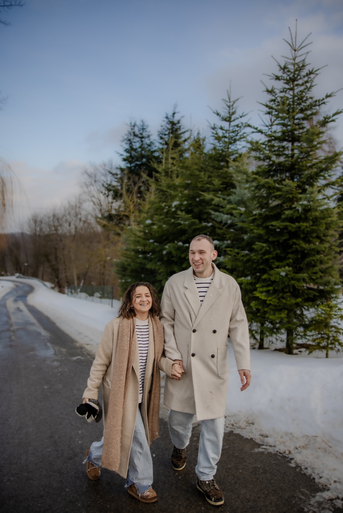 Uśmiechnięta para w zimowych płaszczach spaceruje po ośnieżonej drodze, trzymając się za ręce na tle zielonych drzew iglastych i błękitnego nieba.