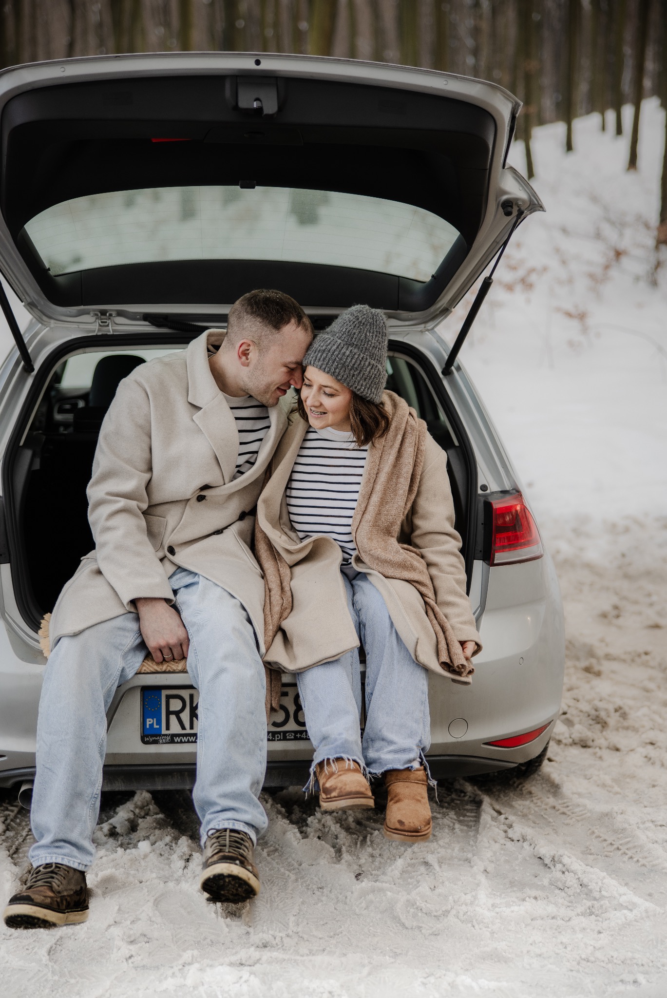 Para w dopasowanych płaszczach siedzi w bagażniku srebrnego auta na tle zimowego lasu. Ona w czapce, oboje w pasiastej odzieży. Widoczne tablice rejestracyjne.
