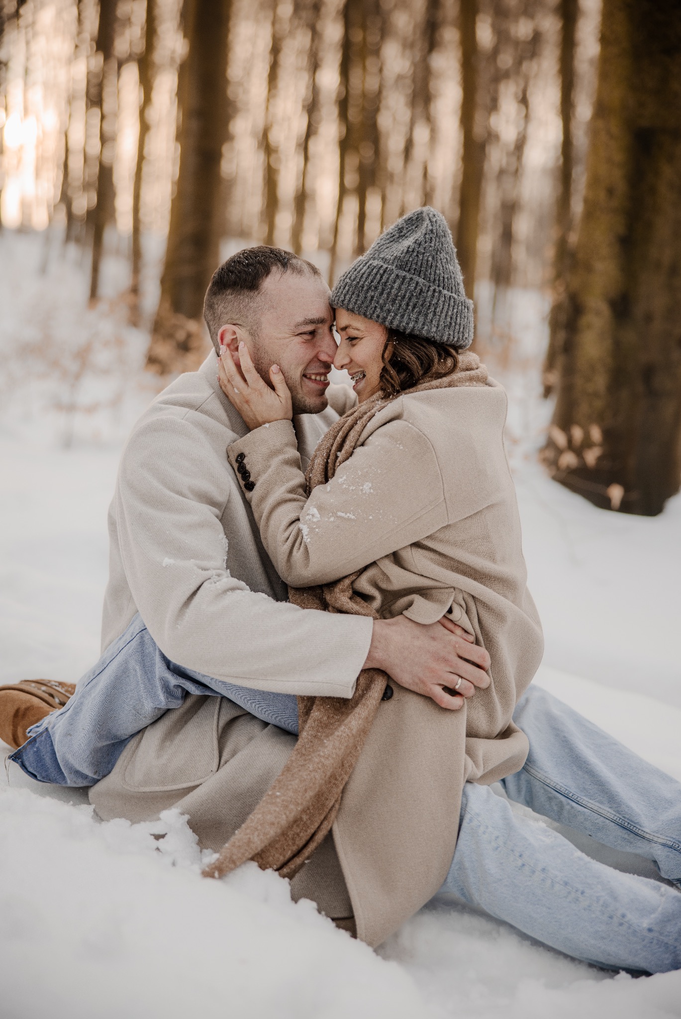 Uśmiechnięta para w zimowej scenerii: mężczyzna obejmuje kobietę w szarym płaszczu i czapce, siedząc na śniegu w lesie. Romantyczna sesja w naturalnym świetle.