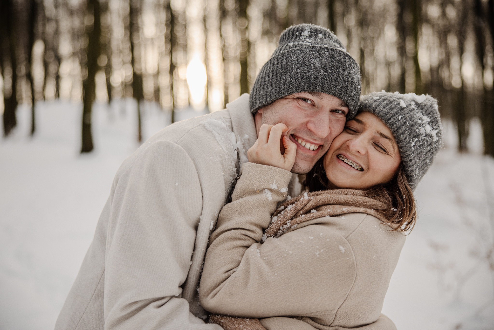Uśmiechnięta para w zimowych ubraniach, obejmująca się na tle ośnieżonego lasu. Dziewczyna ma aparat ortodontyczny. Ujęcie z bliska, ciepłe światło.