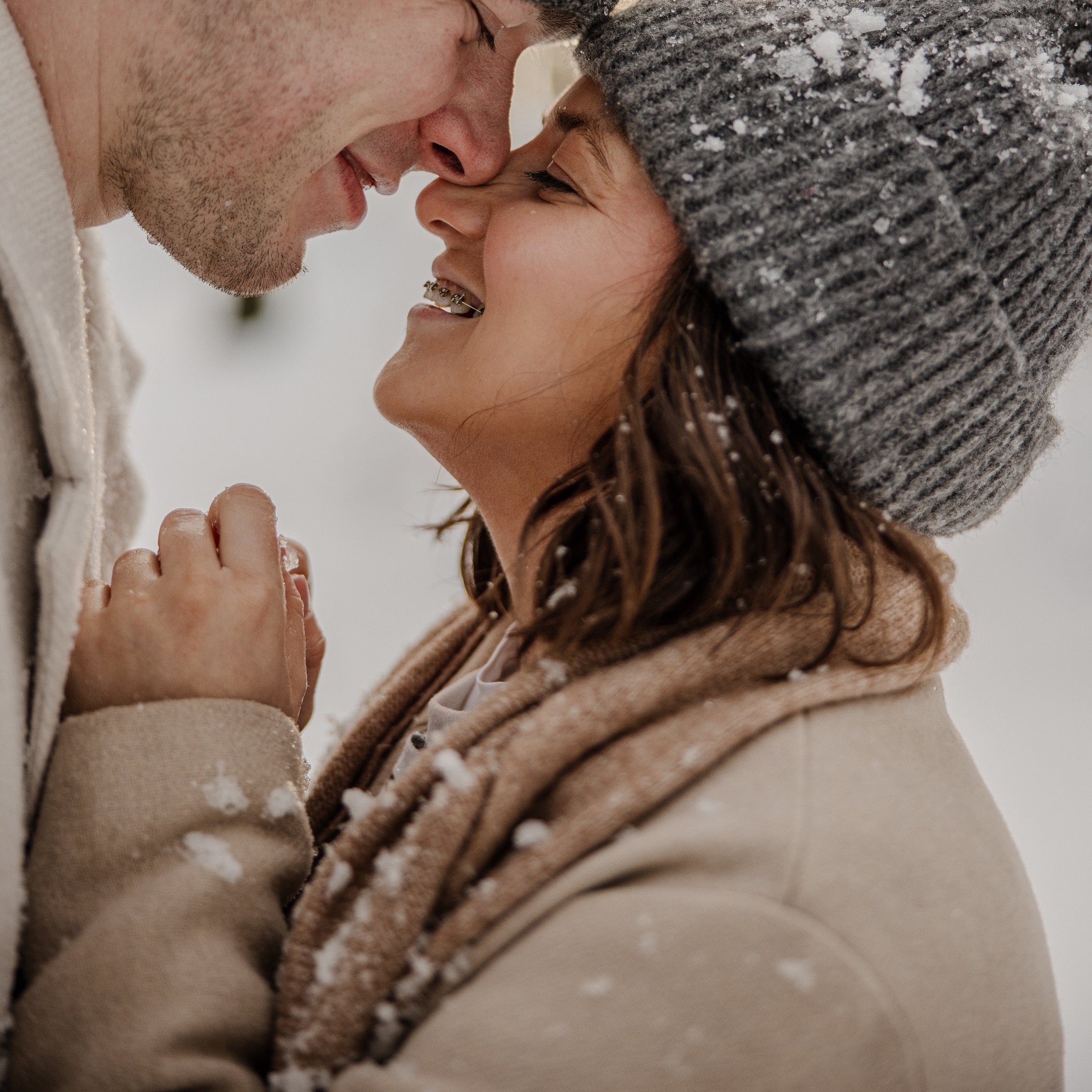 Zimowe zbliżenie na parę: on w jasnym swetrze, ona w czapce i płaszczu, dotykają się nosami, uśmiechając się. Na ubraniach i włosach widoczne płatki śniegu.