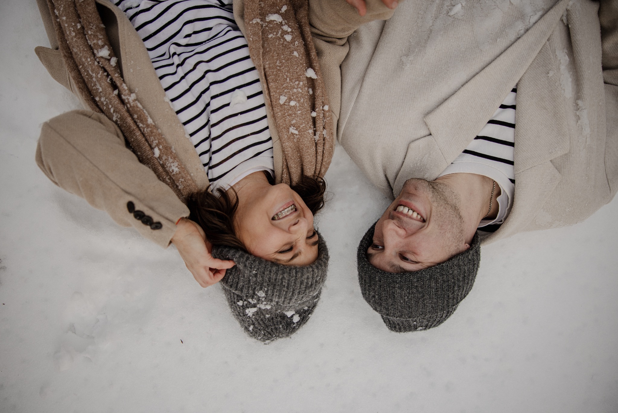 Uśmiechnięta para w zimowych czapkach leży na śniegu, widok z góry. Ona w beżowym płaszczu i bluzce w paski, on w jasnym płaszczu. Śnieg na czapkach.