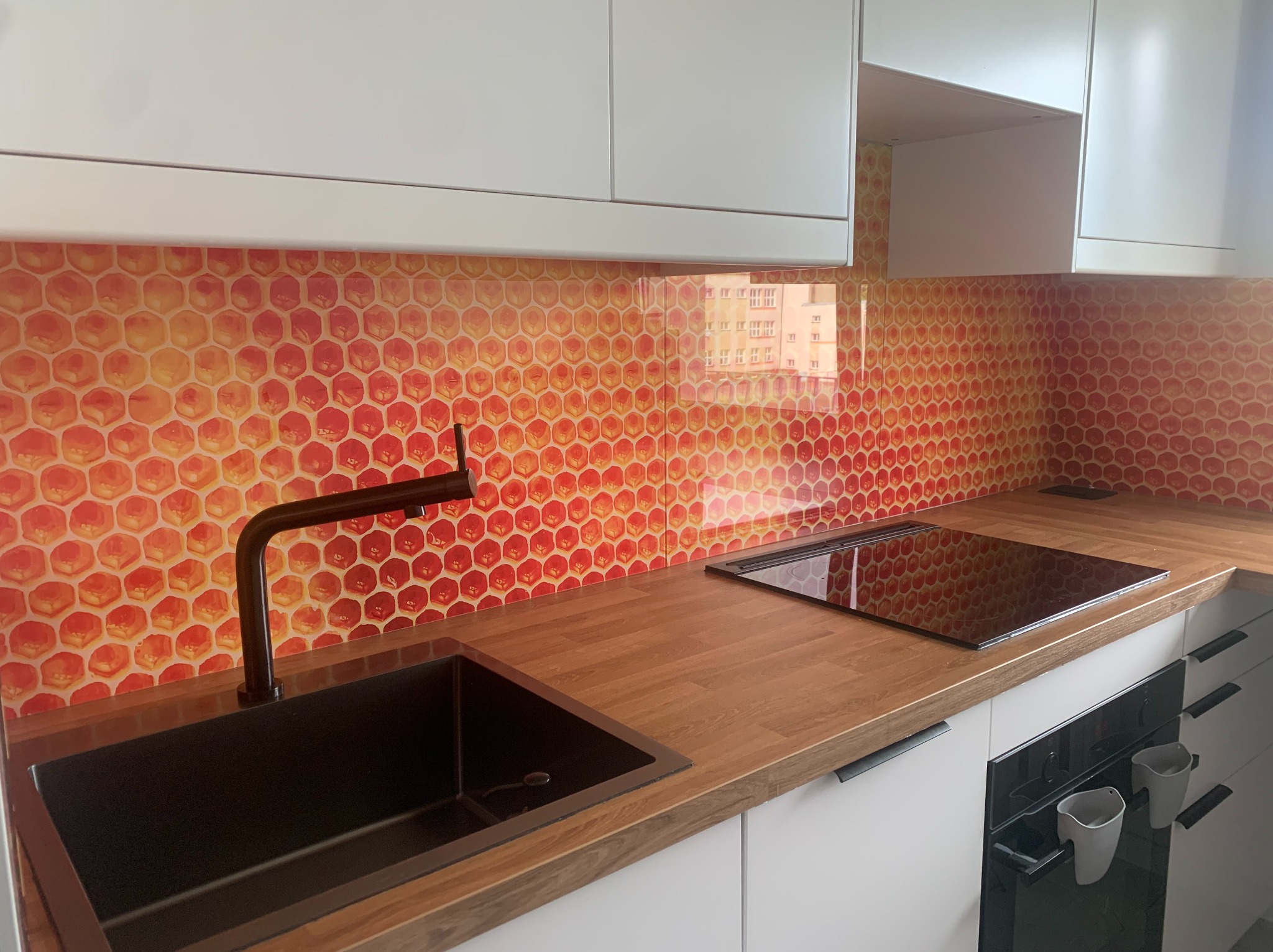 Nowoczesna kuchnia z blatem drewnopodobnym i oryginalną, pomarańczowo-czerwoną mozaiką heksagonalną na ścianie. Czarny zlew i bateria, białe szafki.