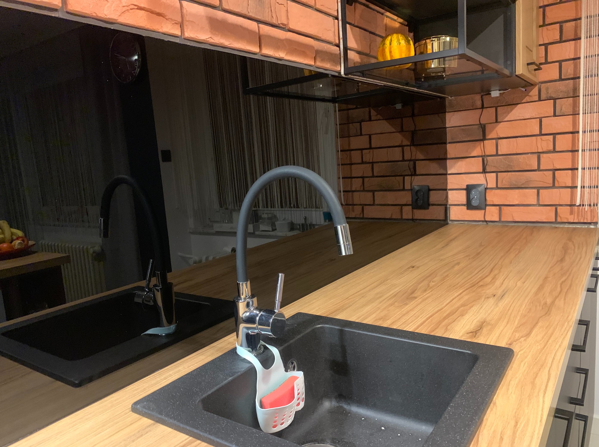 Nowoczesna kuchnia z czarnym, granitowym zlewem i baterią na tle ceglanej ściany. Nad blatem szafka z czarnym szkłem odbijającym wnętrze.