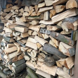 Wynajem sprzętu i urządzeń Marcel Roczyna - Ułożony stos pociętego drewna opałowego, zabezpieczony drewnianą konstrukcją i plandeką. Widoczne różne odcienie drewna i kora. Tło ziemiste.