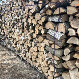 Wynajem sprzętu i urządzeń Marcel Roczyna - Rząd ułożonego drewna kominkowego na palecie, z widocznymi śladami cięcia i oznaczeniami, gotowego do sezonowania lub transportu. Widoczne trociny na podłożu.