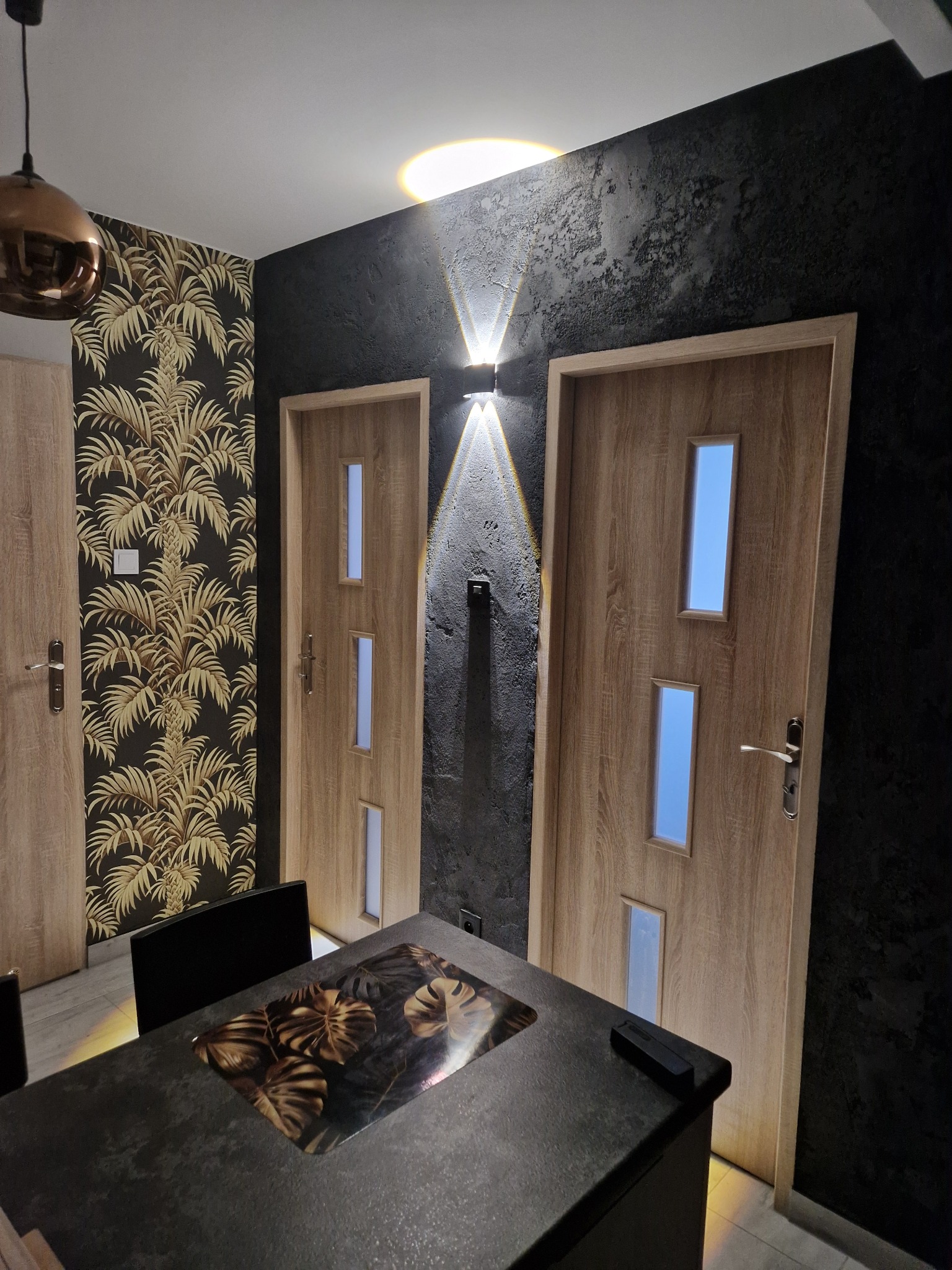 Nowoczesne wnętrze po remoncie: ciemna ściana z fakturą, dwoje drzwi z przeszkleniami, designerska lampa ścienna i stół z blatem imitującym kamień.