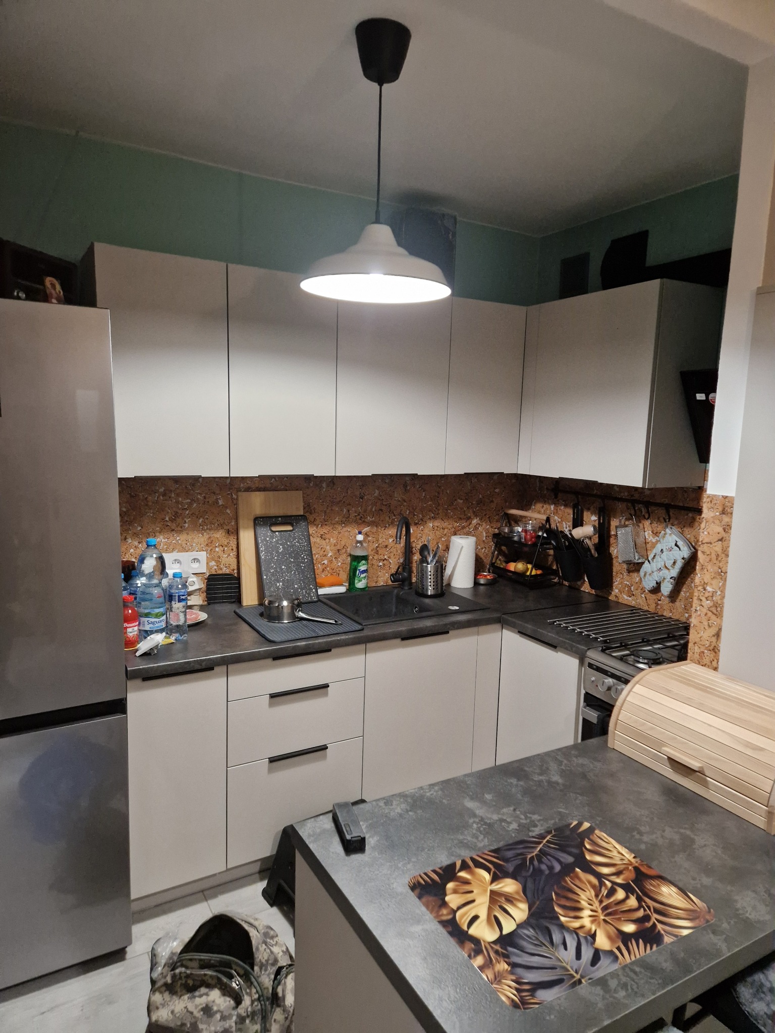 Nowoczesna kuchnia z białymi szafkami, korkowym panelem ściennym i czarnym blatem. Na blacie akcesoria kuchenne i dekoracyjny podkładka pod talerze.