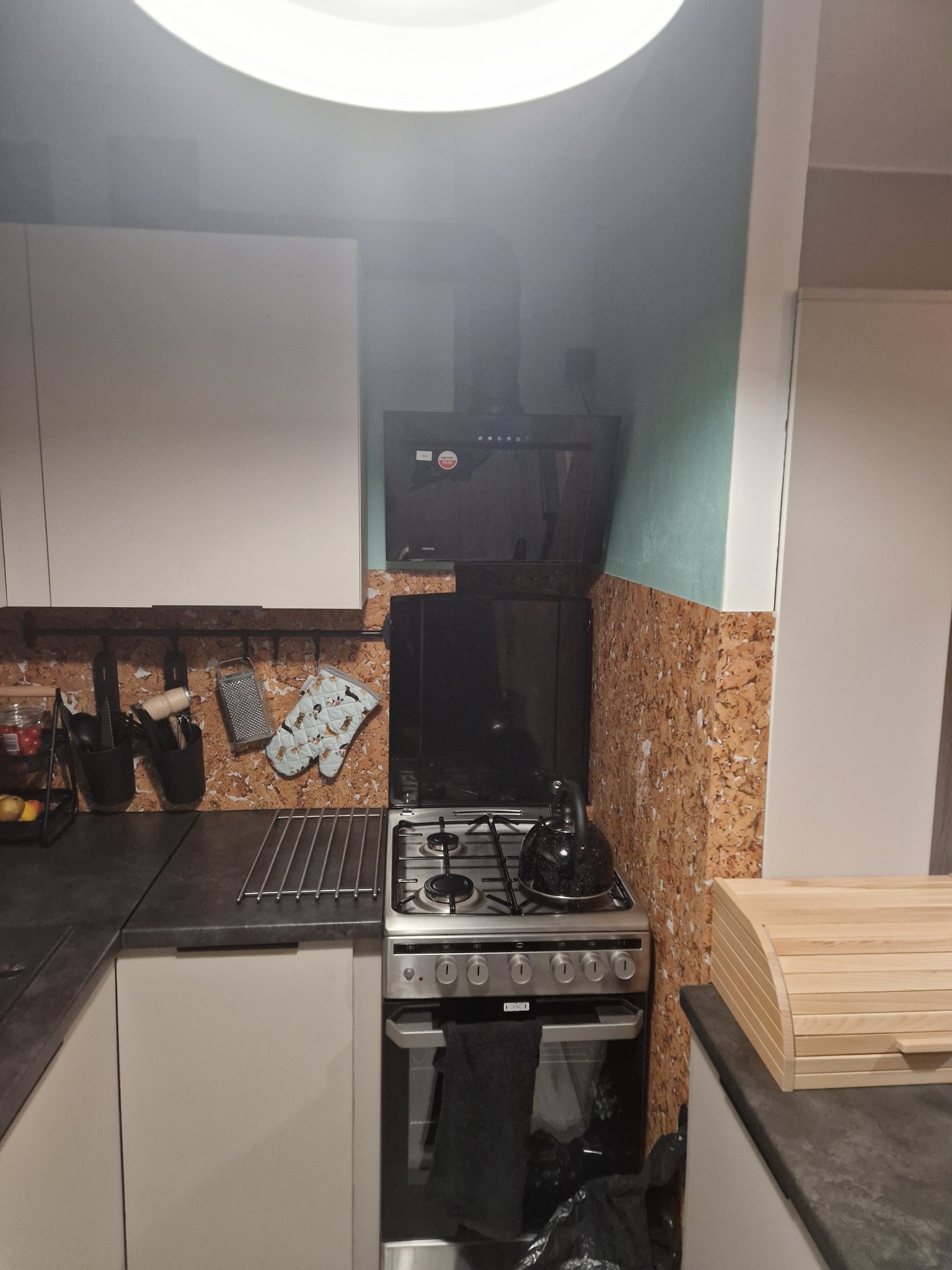 Kuchnia po remoncie: białe szafki, korkowa ściana, nowoczesny okap i kuchenka gazowa. Na blacie chlebak i czajnik. Widoczne akcesoria kuchenne.