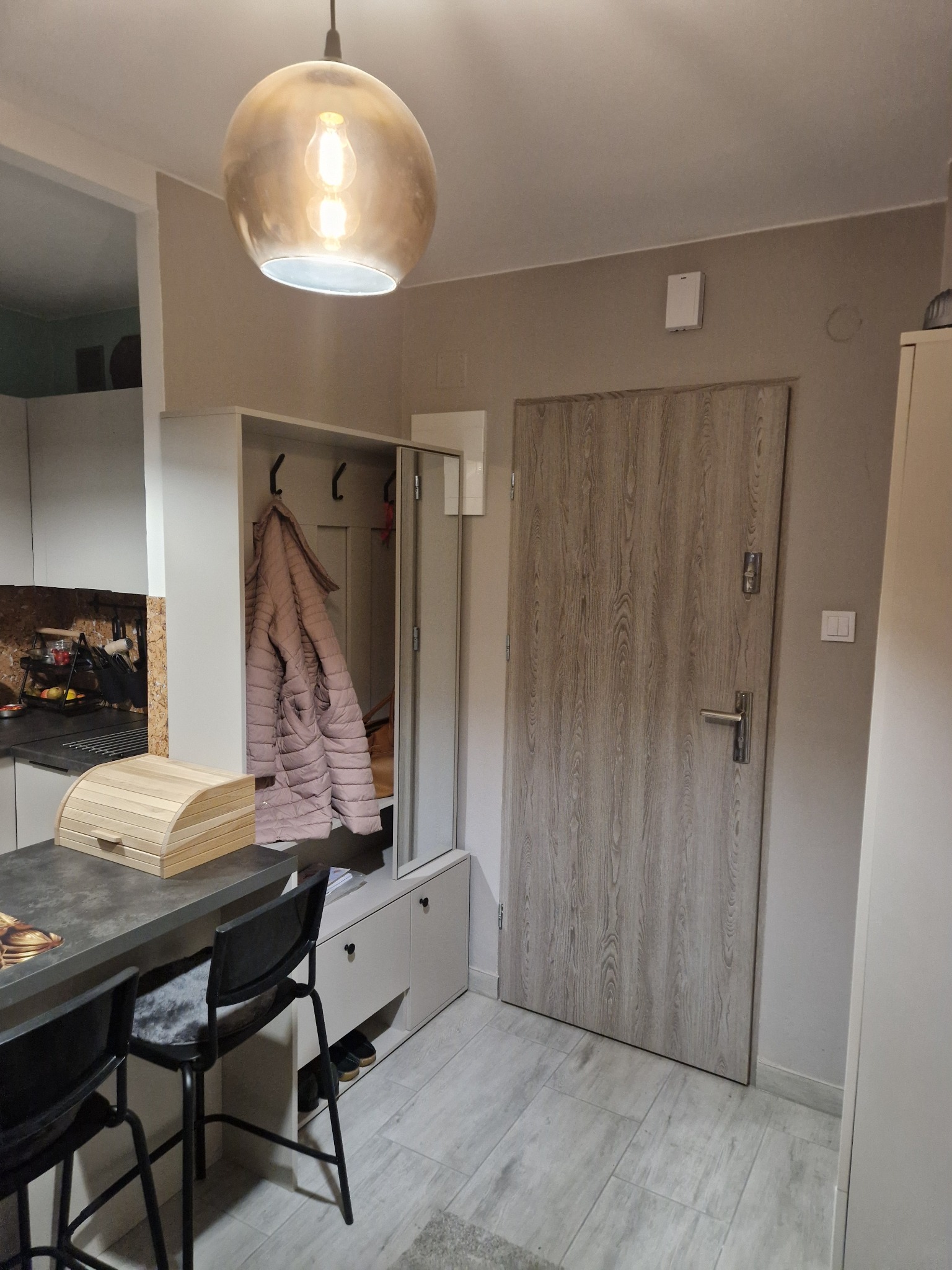 Jasne wnętrze mieszkania z szafą wnękową, wieszakami i lustrem. Widoczna lampa wisząca, stół z krzesłami i drzwi wejściowe. Neutralna kolorystyka ścian i podłogi.