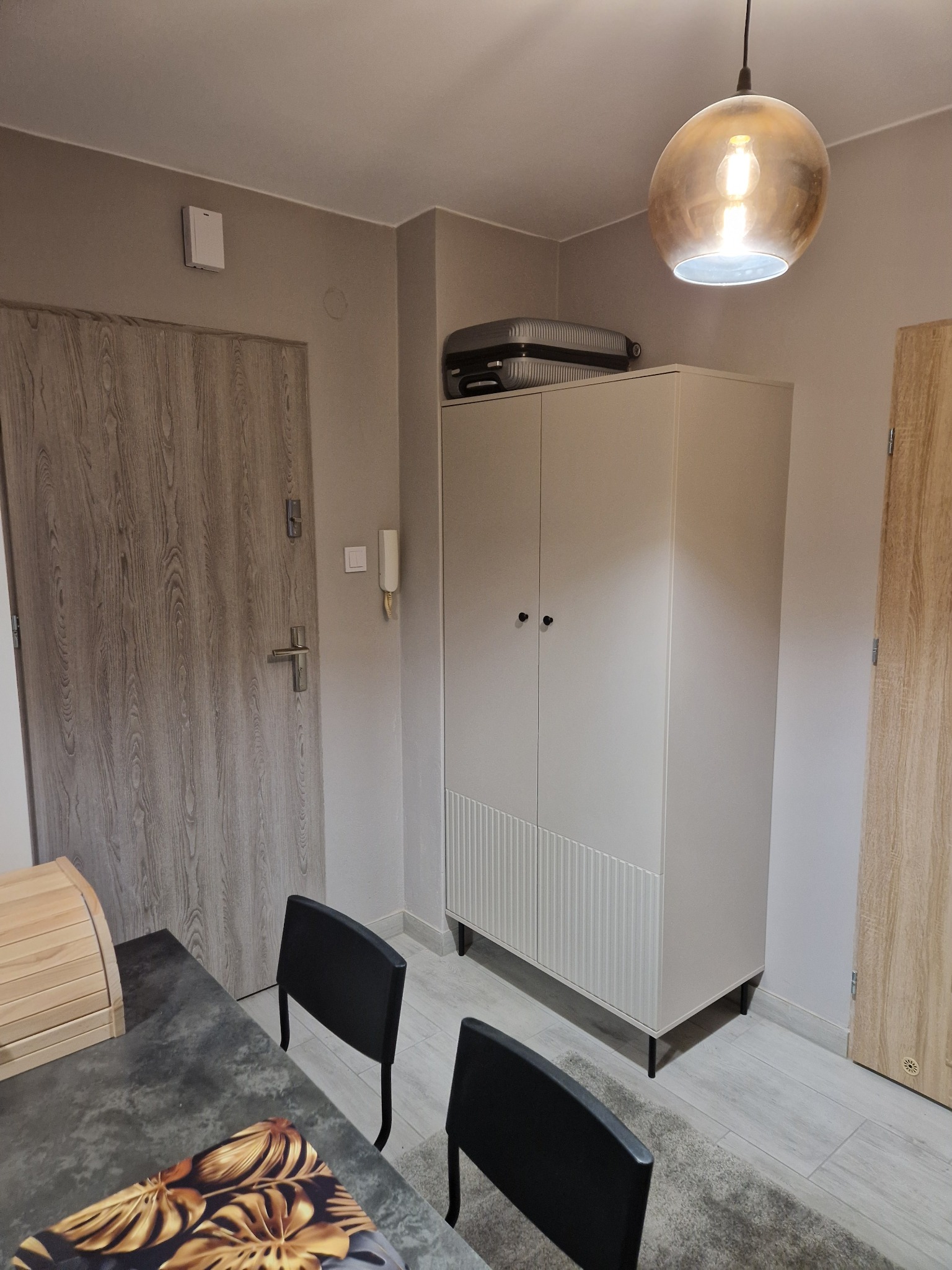 Jasne wnętrze mieszkania z szafą, drzwiami i lampą wiszącą. Na szafie leży szara walizka. Widoczne krzesła i fragment stołu z dekoracyjną podkładką.