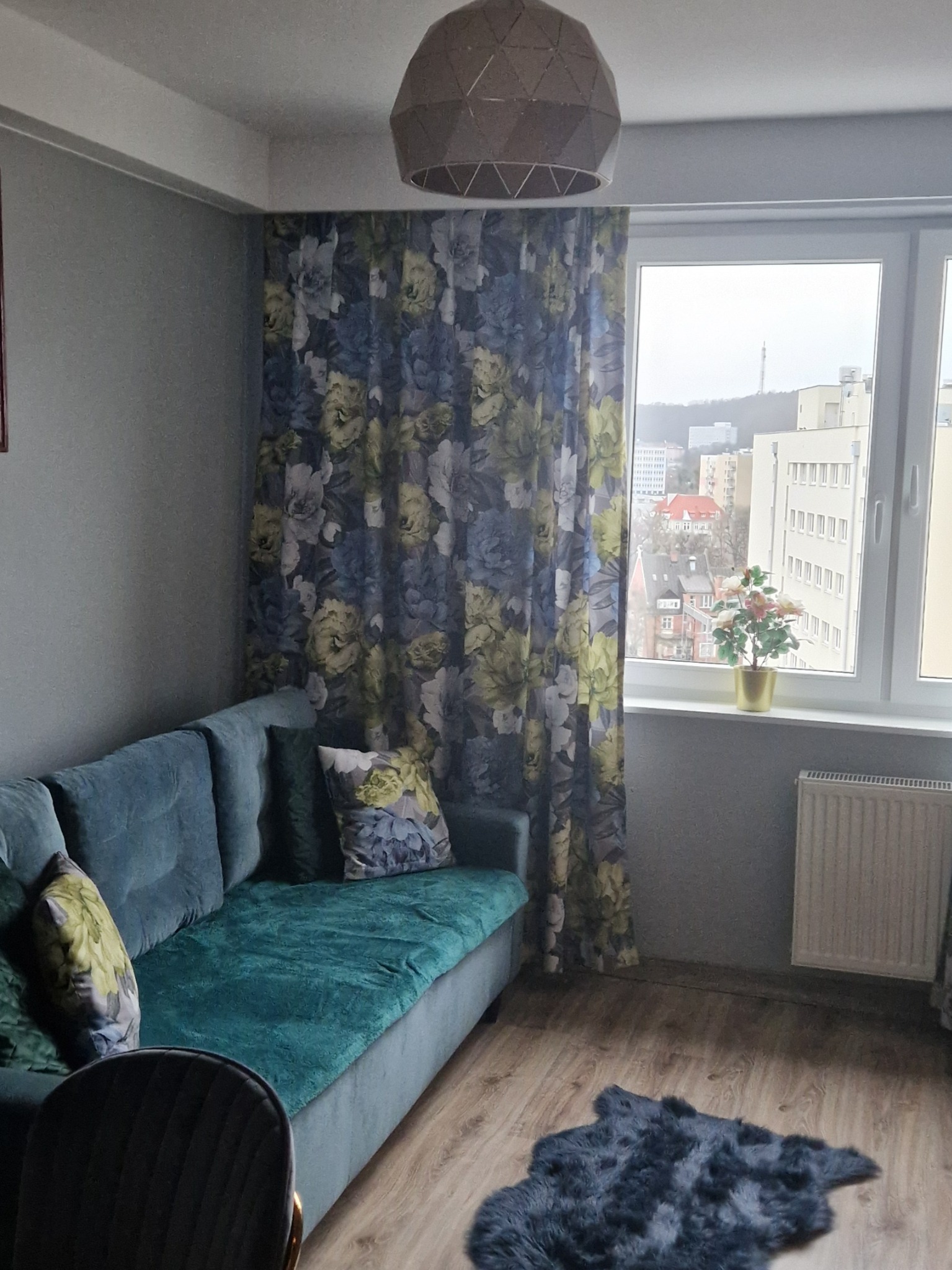 Stylowy salon z turkusową sofą, wzorzystymi zasłonami i widokiem na miasto za oknem. Na podłodze leży ciemny dywanik, a całość oświetla designerska lampa.
