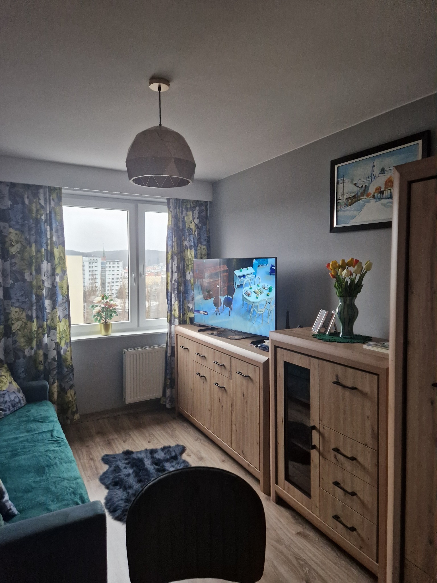 Jasne drewniane meble w pokoju z szarą ścianą, kwiatami i telewizorem. Widok z okna na miasto. Dywanik na podłodze. Lampa sufitowa w geometrycznym stylu.