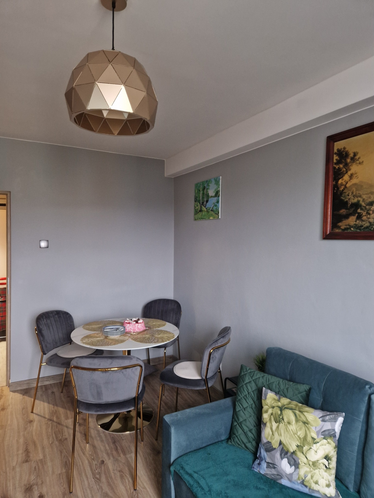Szare wnętrze pokoju z okrągłym stołem, krzesłami na złotych nogach, turkusową sofą i geometryczną lampą. Na ścianach wiszą obrazy.