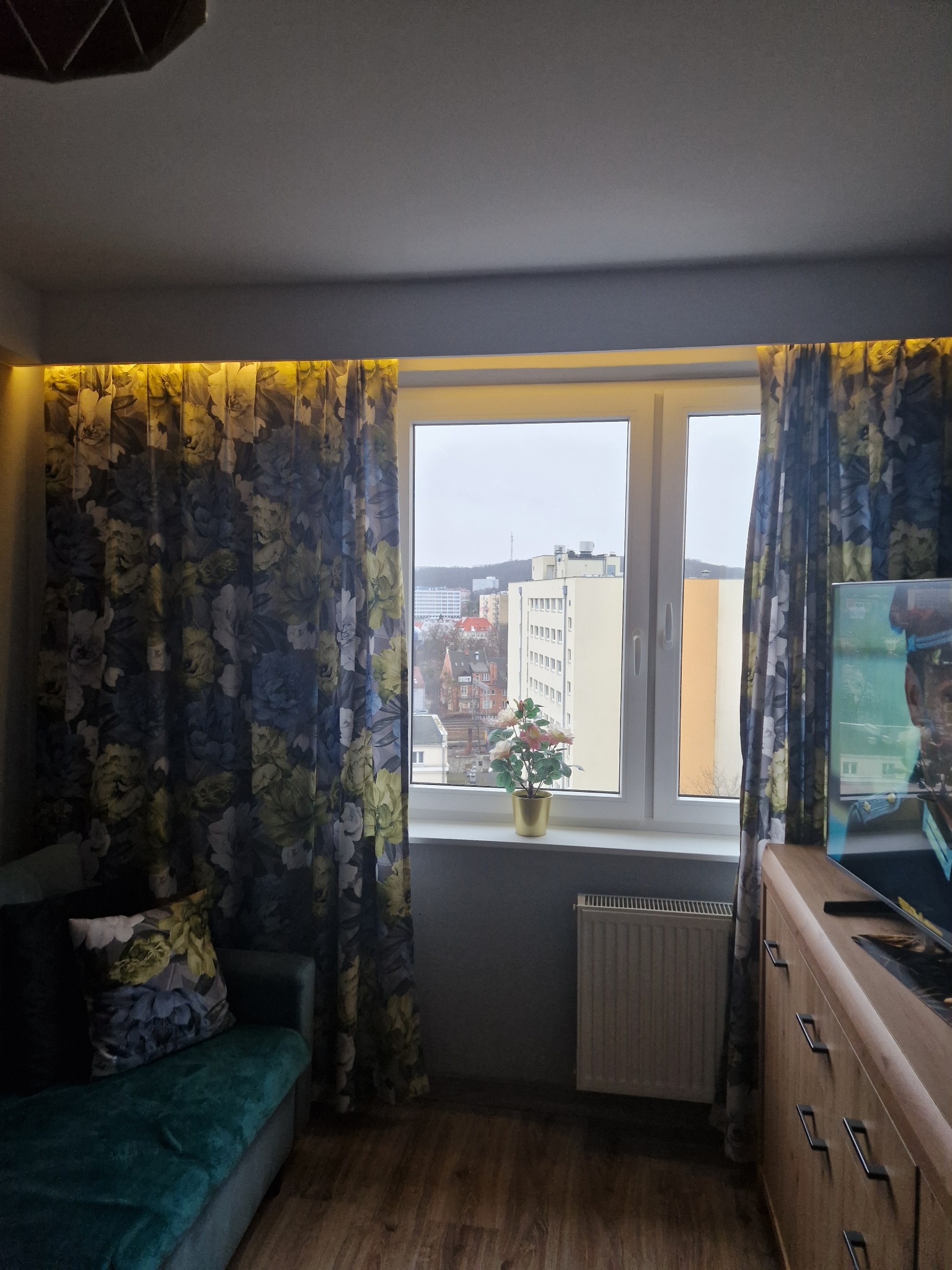 Pokój po remoncie: okno z kwiatami, zasłony w kwiaty z podświetleniem LED, sofa z poduszką w kwiaty, drewniana komoda. Widok na miasto za oknem.