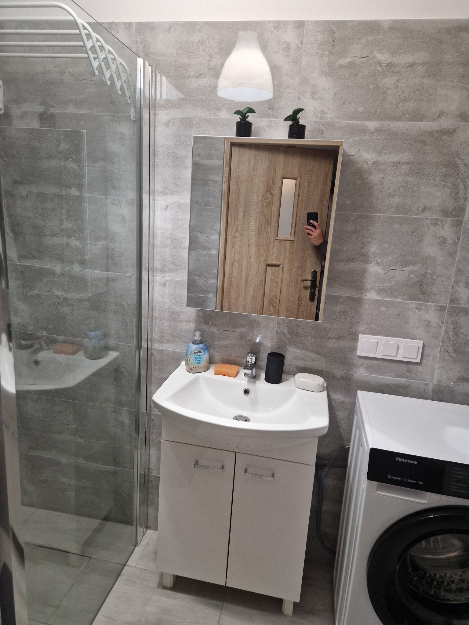 Szara łazienka po remoncie z kabiną prysznicową, umywalką z szafką, pralką Hisense i lustrem, w którym odbija się drzwi. Drobne rośliny na szafce.