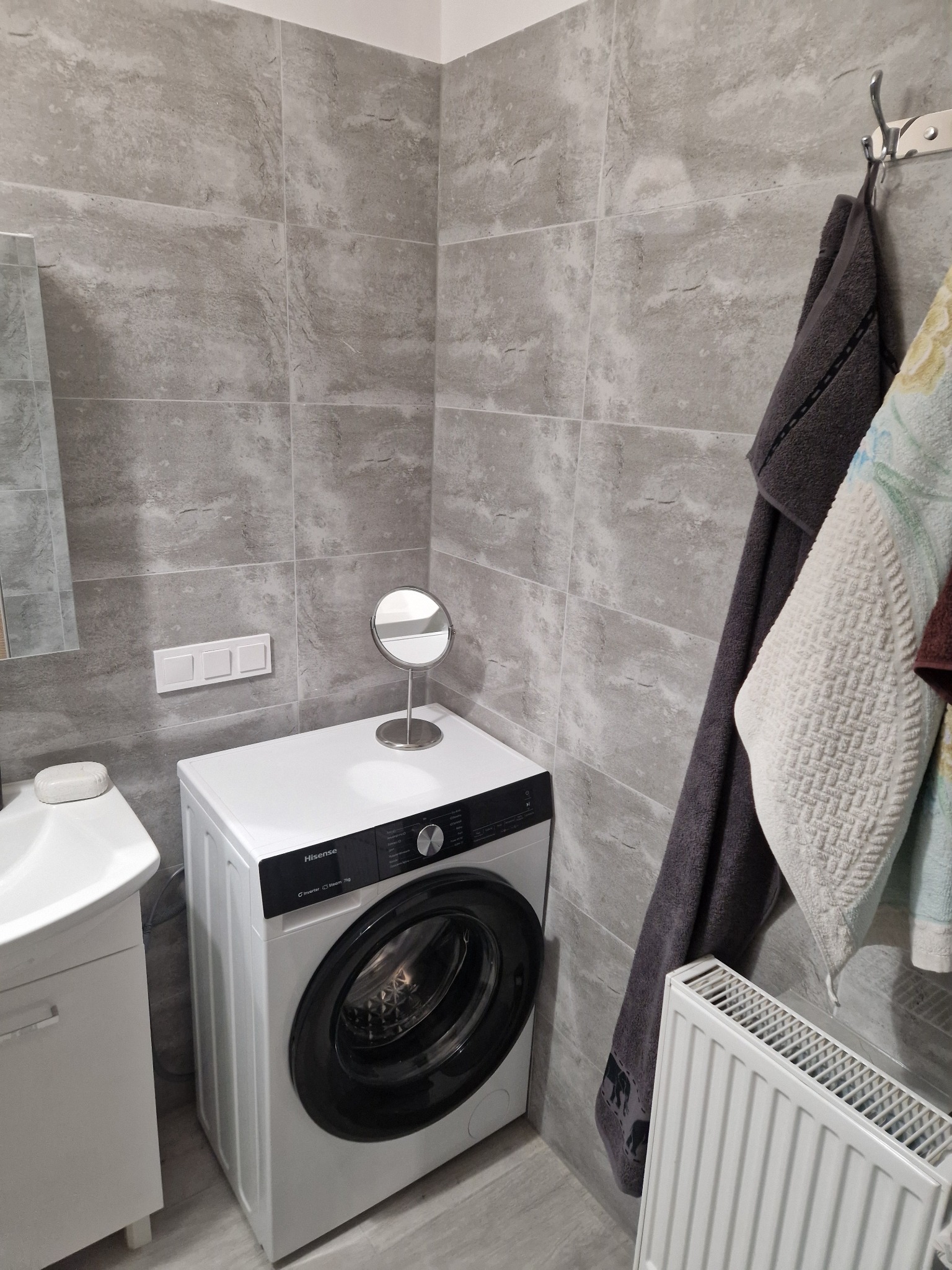 Szara łazienka po remoncie: pralka Hisense, umywalka z szafką, grzejnik, lustro na blacie pralki, ręczniki na wieszaku, płytki imitujące kamień.