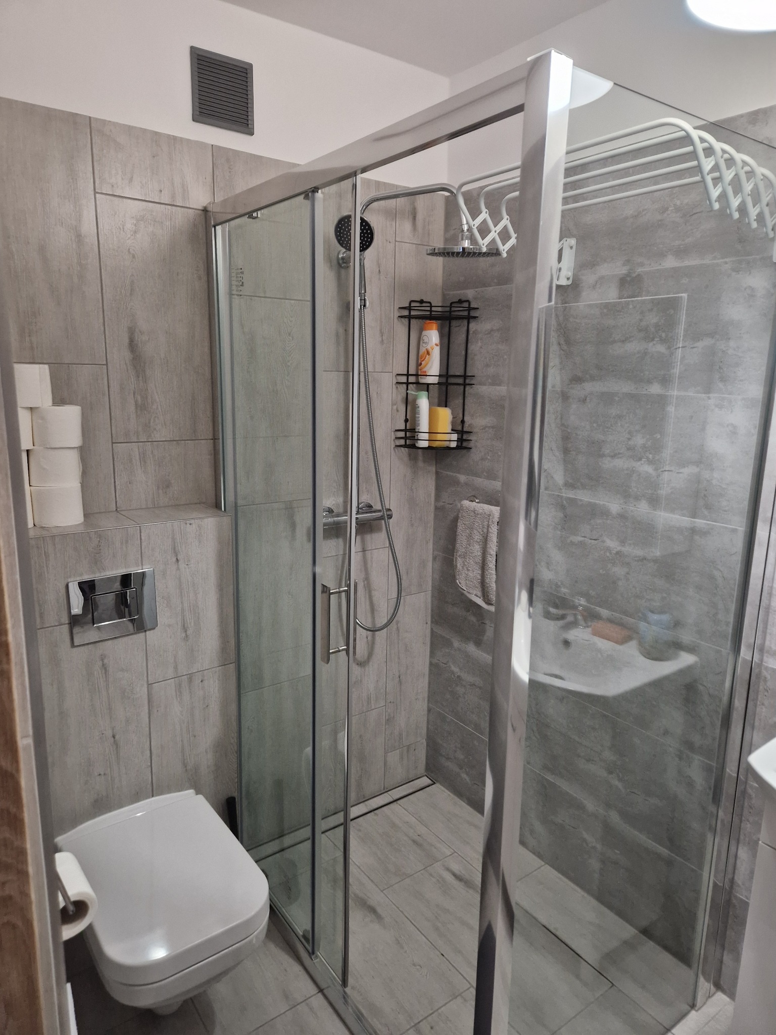 Nowoczesna łazienka z kabiną prysznicową typu walk-in, wykończona szarymi płytkami imitującymi drewno. Widoczna toaleta i półka z kosmetykami pod prysznicem.