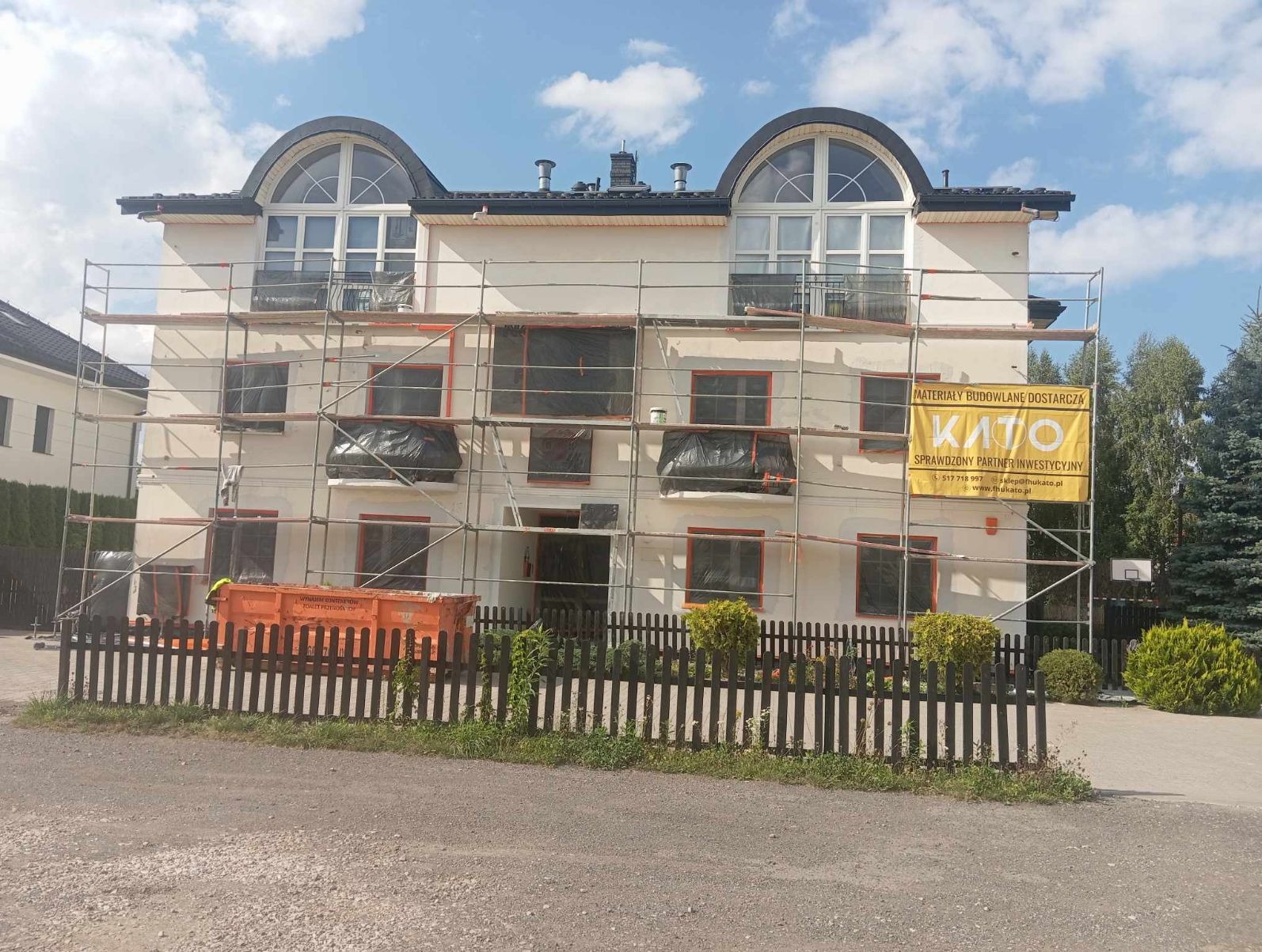 Dwupiętrowy budynek w trakcie prac elewacyjnych, z rusztowaniami i oknami zabezpieczonymi folią. Widoczny kontener na odpady i baner reklamowy na rusztowaniu.