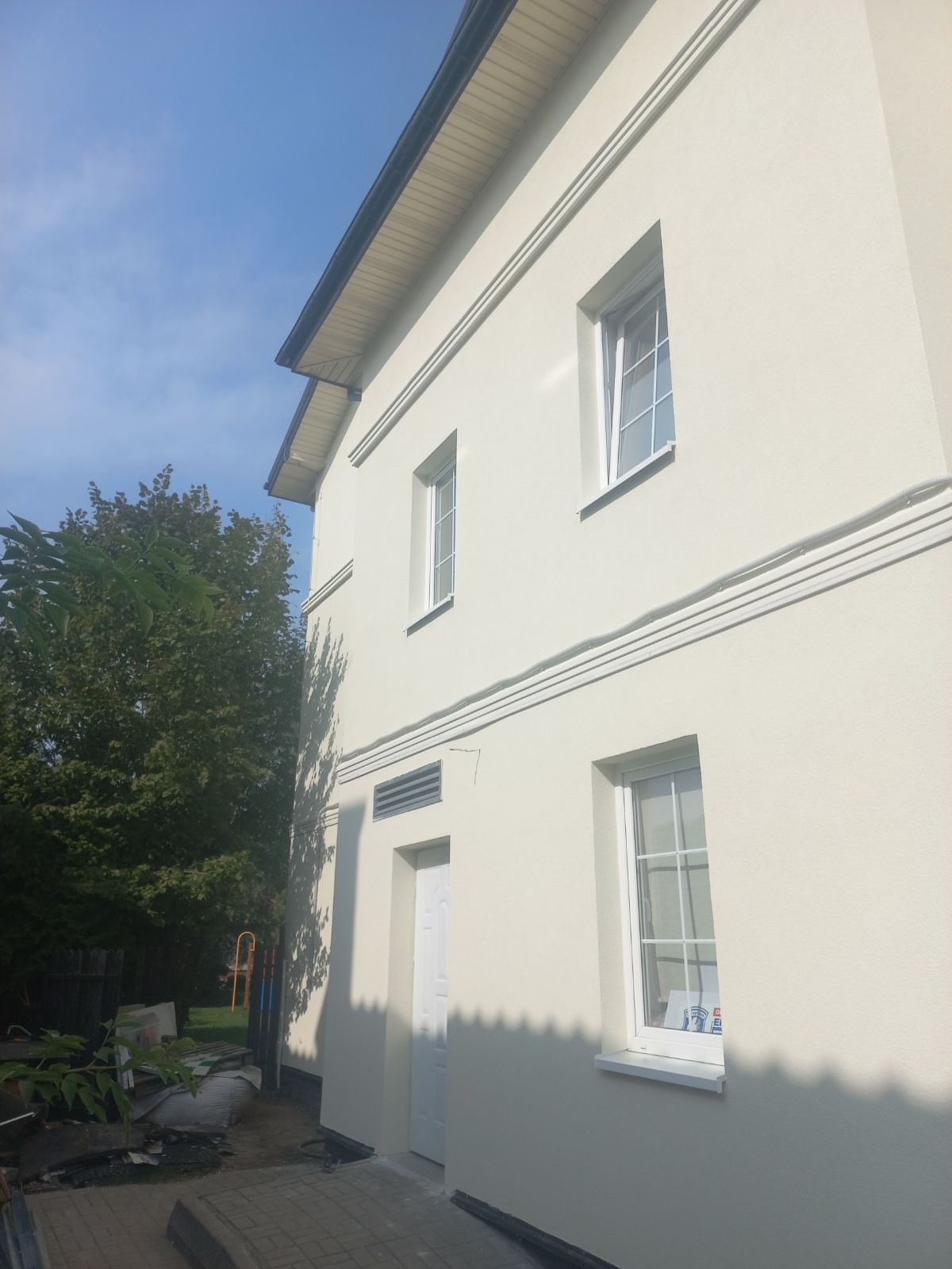 Jasna elewacja budynku po renowacji, widok z dołu, z oknami i elementami dekoracyjnymi. Widoczne przewody na elewacji. Obok drzewo i fragment nieba.