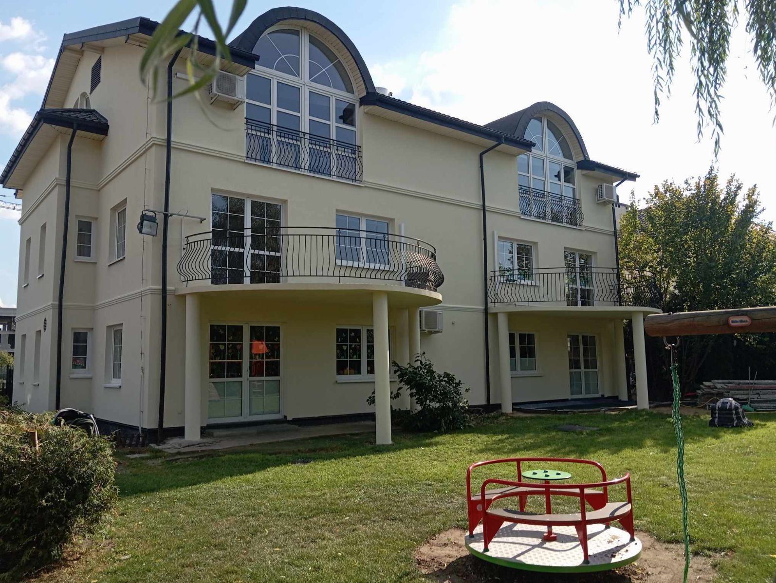 Dwupiętrowy budynek z kremową elewacją, balkonami z czarnymi balustradami i dużymi oknami. Na pierwszym planie trawnik z karuzelą. Widoczne klimatyzatory na ścianie.