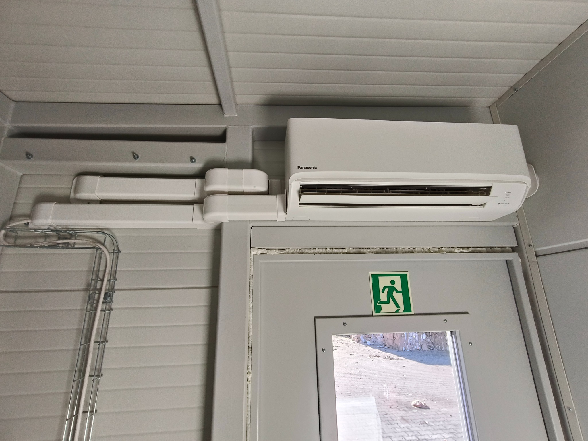 Zamontowany klimatyzator Panasonic w pomieszczeniu z szarą ścianą panelową i instalacją elektryczną w korytku. Widoczny znak ewakuacyjny na drzwiach.
