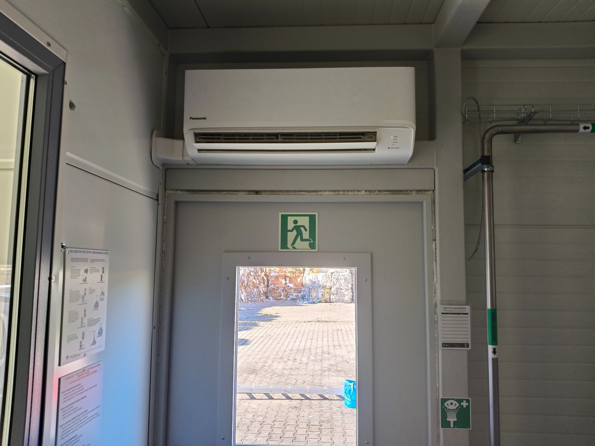 Biały klimatyzator Panasonic zamontowany nad szarymi drzwiami z szybą, widok na zewnątrz. Tabliczki informacyjne na ścianie, rura z oznaczeniem kierunku.