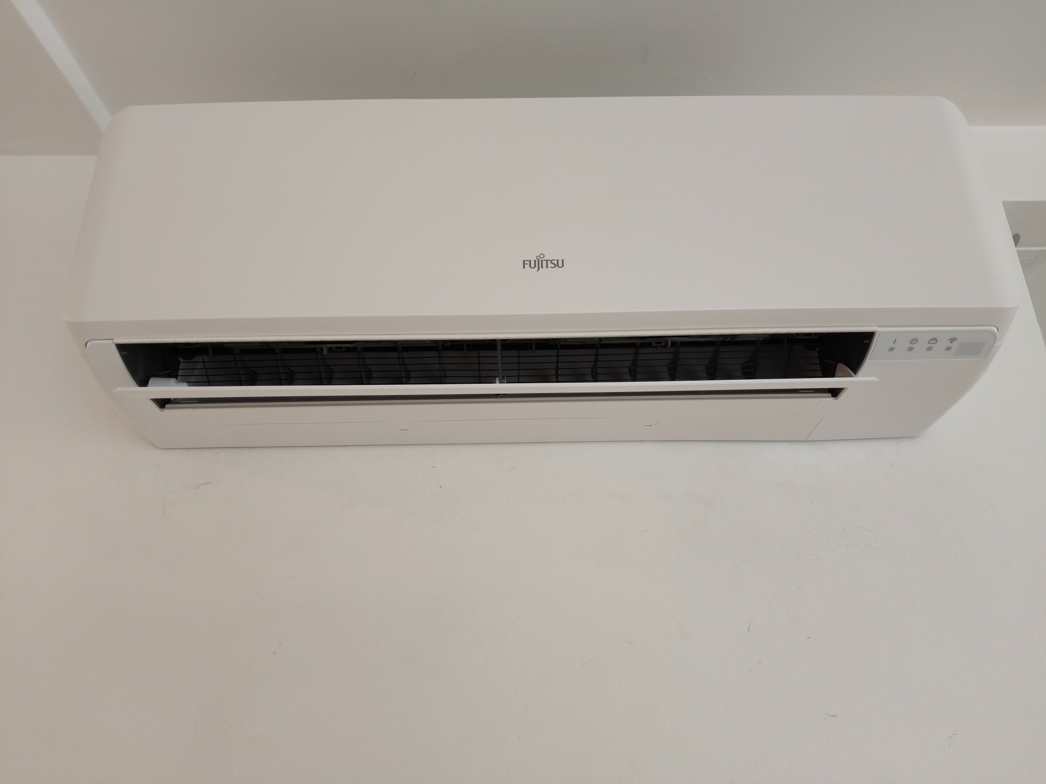 Biała jednostka klimatyzacji Fujitsu zamontowana na jasnej ścianie. Widoczne logo i panel sterowania. Minimalistyczny design, czyste linie.