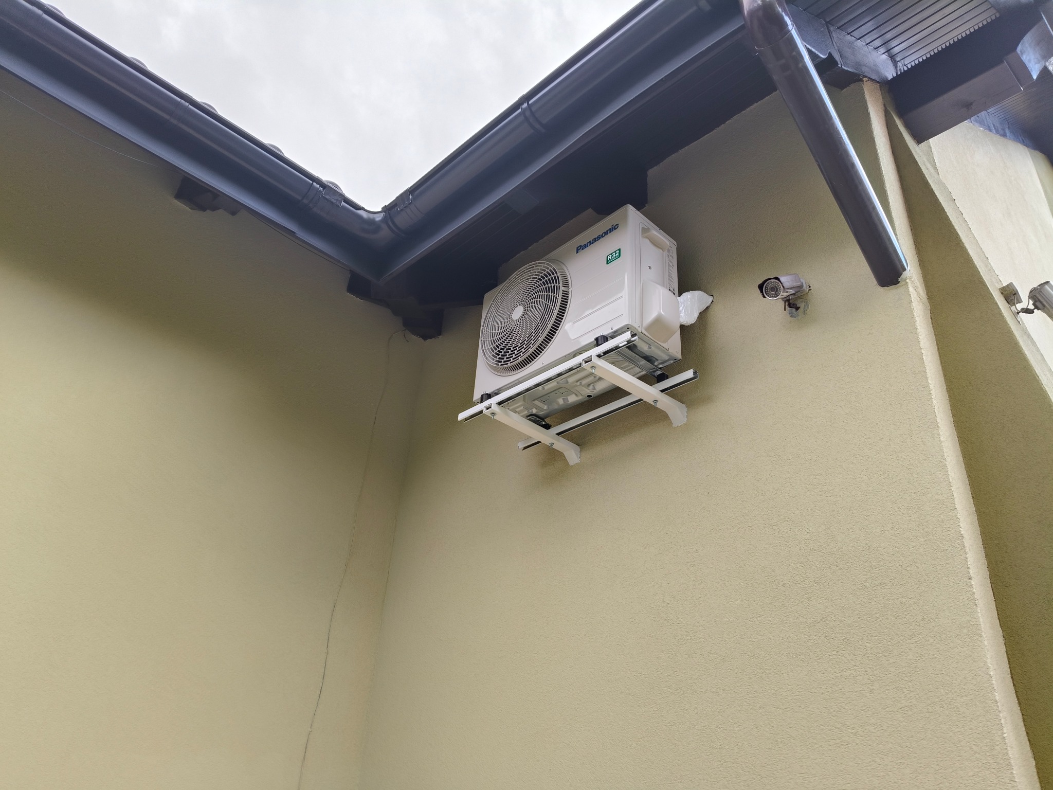 Biała jednostka zewnętrzna klimatyzacji Panasonic R32 zamontowana na kremowej ścianie budynku, widoczna kamera monitoringu i brązowa rynna dachowa.