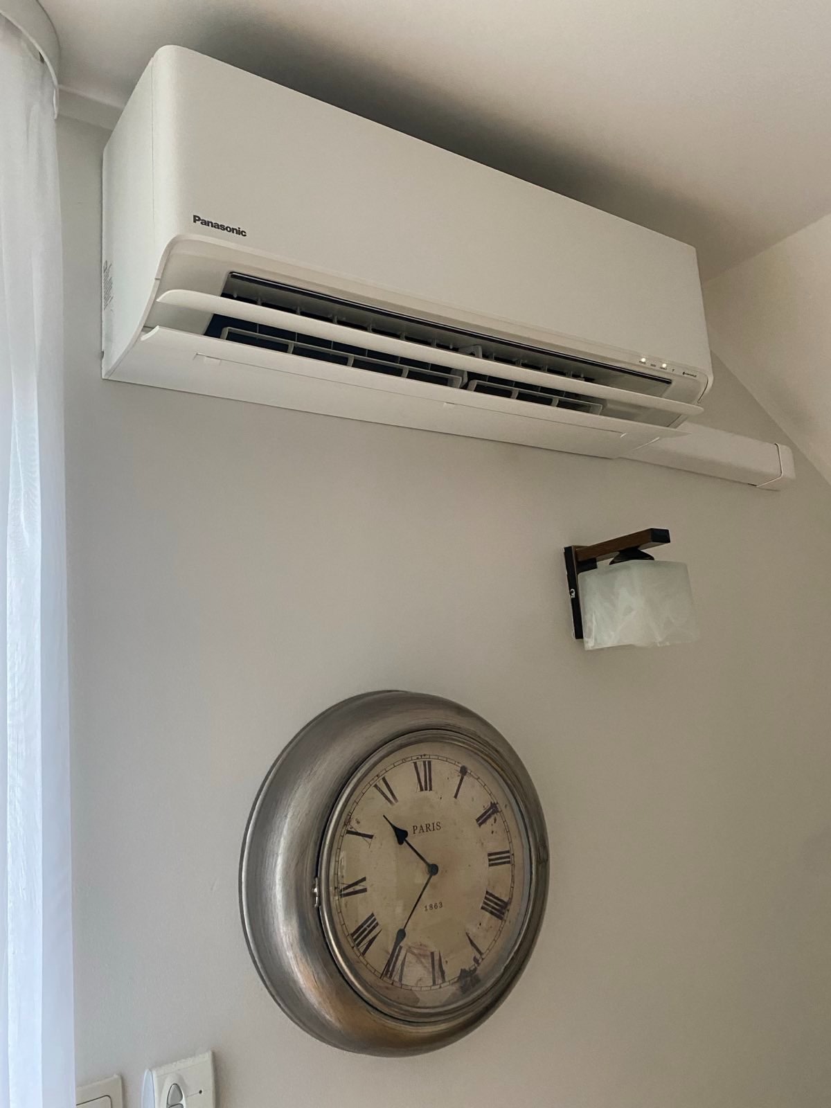 Biała klimatyzacja Panasonic na ścianie w pokoju, obok zegar w stylu vintage i lampka. Widok z dołu, oświetlenie dzienne.