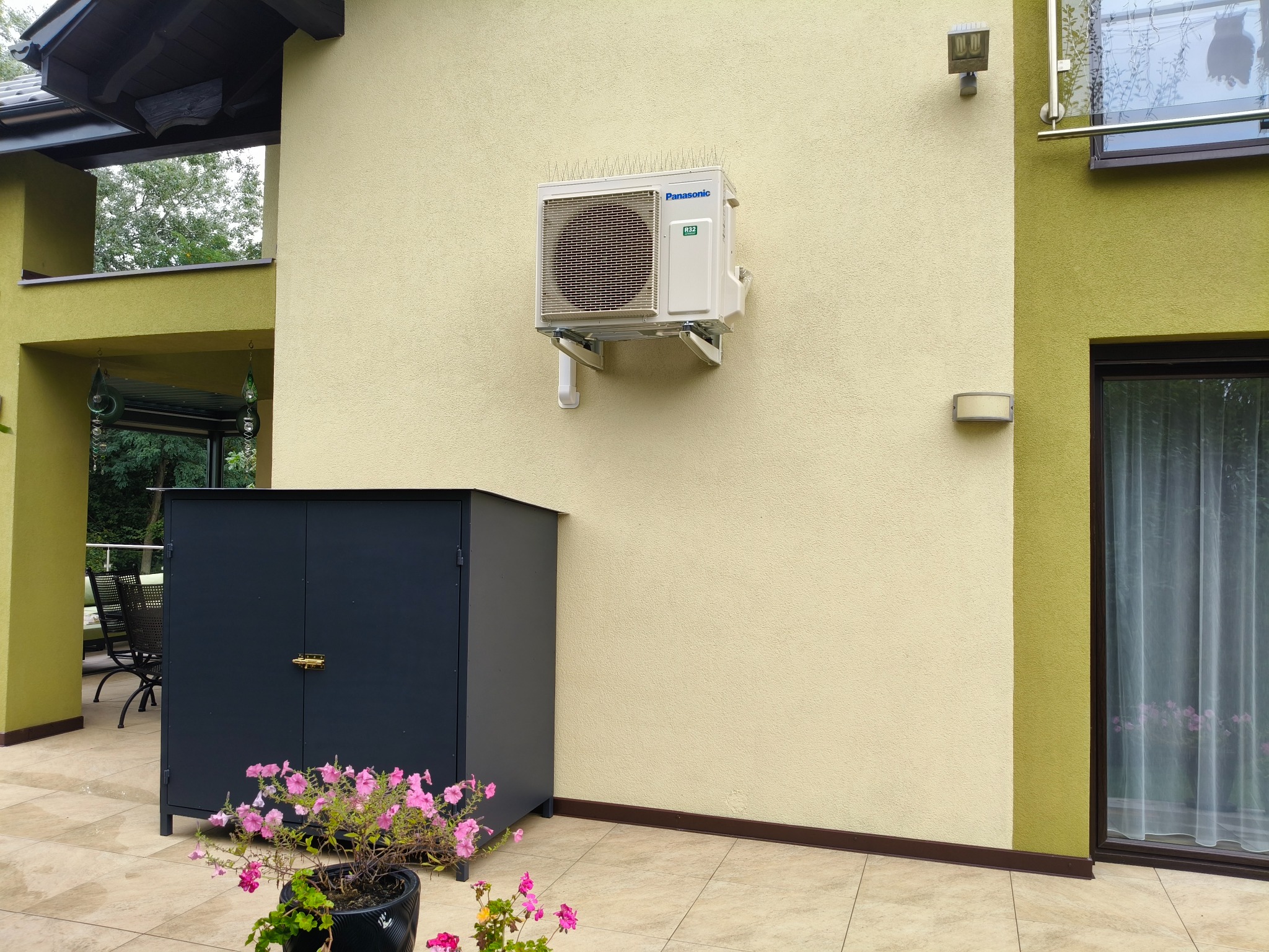 Zamontowany na elewacji budynku klimatyzator Panasonic z kolcami przeciw ptakom. Obok szafka narzędziowa i kwiaty w doniczce na tarasie.