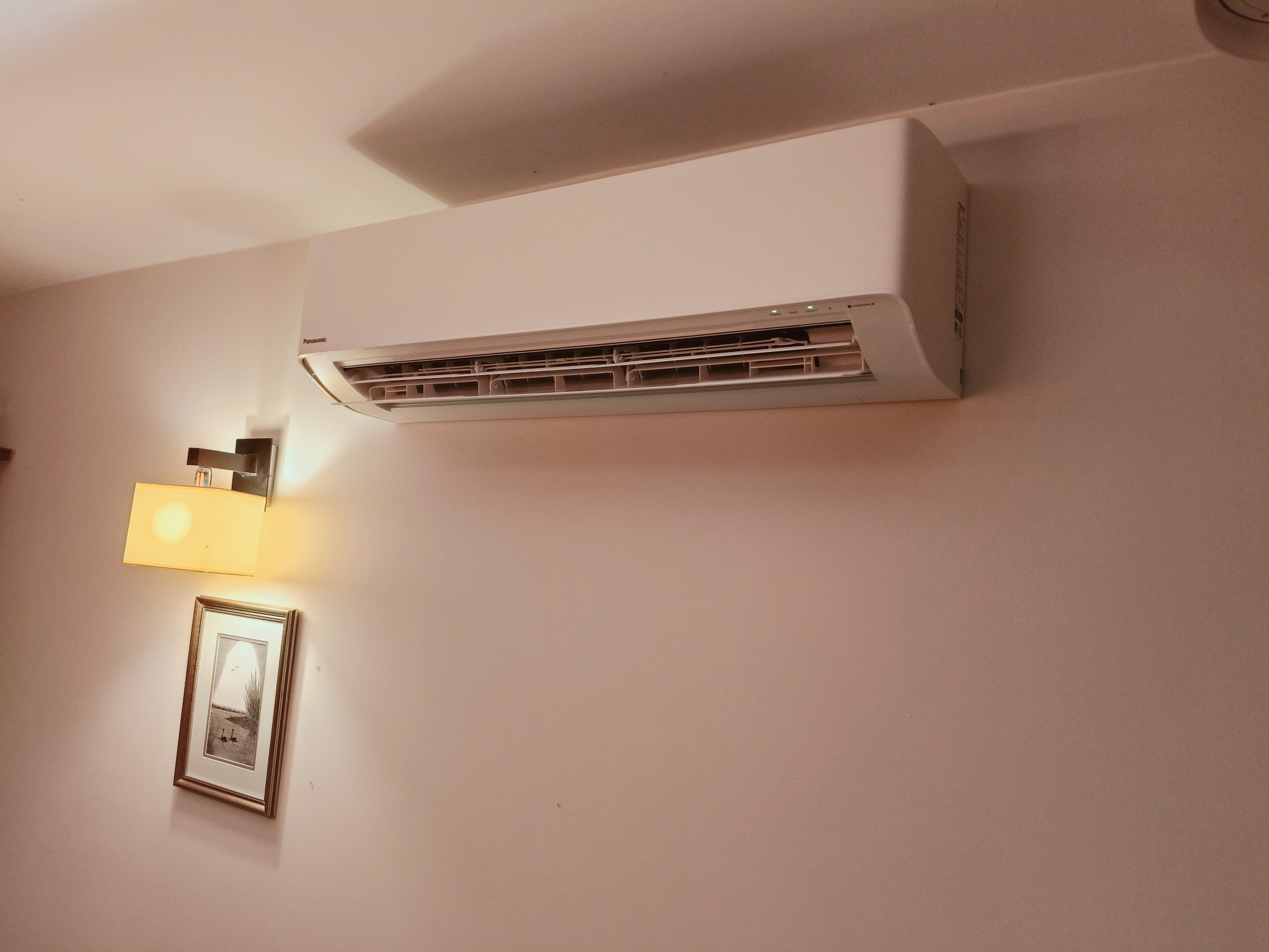 Biały klimatyzator Panasonic zamontowany na ścianie w pokoju, obok lampki i obrazu w ramce. Widoczne diody sygnalizacyjne na urządzeniu.