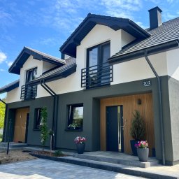 Nowoczesny bliźniak z ciemną elewacją i drewnianymi akcentami w Olsztynie. Widoczne balkony z czarną balustradą, zadbany teren wokół budynku i błękitne niebo.