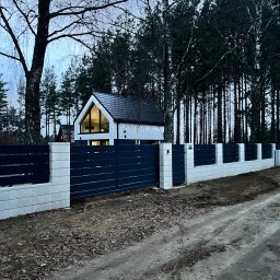 Nowoczesny dom jednorodzinny z ciemną stolarką okienną i ogrodzeniem z betonowych bloczków, otoczony lasem. Ujęcie z poziomu gruntu, widoczna droga gruntowa.