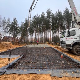 BUD-KOR Grzegorz Korgul - Zbrojenie fundamentu pod dom jednorodzinny, częściowo zalane betonem, z widocznym podajnikiem betonu Bosta-Beton i pracownikami w tle. Teren budowy na obrzeżach lasu.