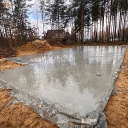 Wylana, mokra płyta fundamentowa na budowie domu jednorodzinnego, zabezpieczona folią. W tle las i częściowo widoczny budynek w stanie surowym.